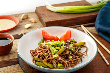 Soba with mushrooms, green beans and sesame seeds in a plate on a stand on a wooden table near chopsticks and ingredients.