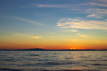 Scenic sunset on lake in dusk with blue clear sky and cirrus clouds, gulls on stone in calm waves under sundown blazing