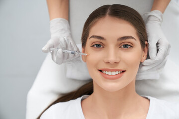Top view portrait of smiling beautiful young woman in white shirt doing procedure for improvements face skin. Concept of special cosmetics injection for anti-aging in beauty salon. - obrazy, fototapety, plakaty