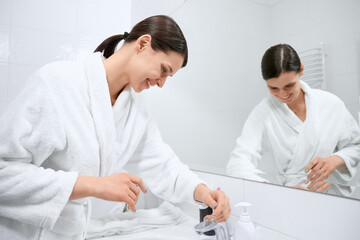 Side view of smiling young brunette woman in white robe washing hands in bathroom near mirror. Concept of water procedure at home or hotel during quarantine. 