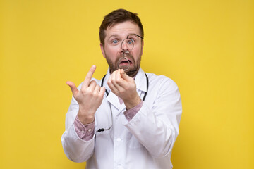 Upset, surprised doctor has taken off glasses and points at them with a foolish expression on face. Yellow background.