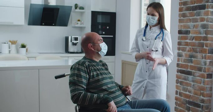 Young woman nurse explaining information to man patient in wheelchair while talking together at home. Epidemic and virus concept. High quality 4k footage