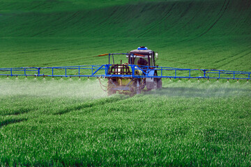 A tractor with attachments performs agricultural work in a green field. Business in industrial agriculture, mechanized processing on the farm. Selective focus, natural background.