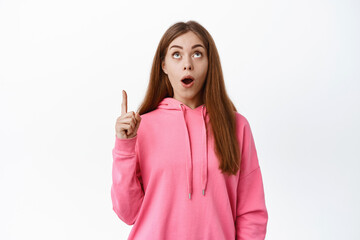 Image of amazed young woman checking out awesome promo deal, gasping and pointing finger up at copy space on top, standing over white background