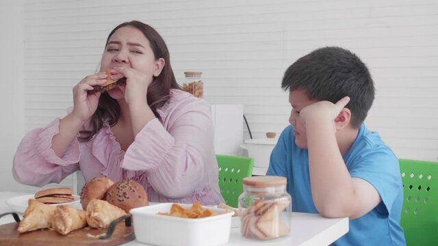 Asian family relationship of son upset with mother fat eating bread in the kitchen at home, mom hungry and eating bun with unhealthy food, two people, woman overweight, health care concept.