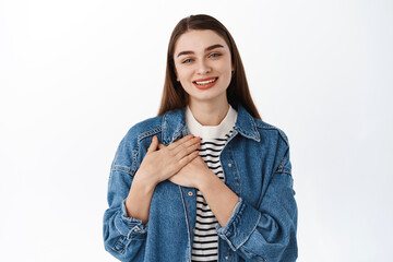 In my heart. Smiling young woman holding hands on chest and looking caring and tender at camera, feeling touched or flattered, heartfelt gaze, standing over white background