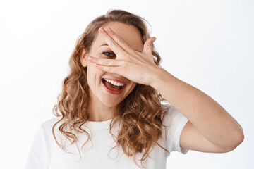 Carefree beautiful woman look through fingers, covering eyes with hand and smiling happy, standing in t-shirt against white background