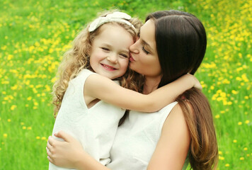 Portrait of little child daughter kissing her happy smiling mother on the grass in a summer day