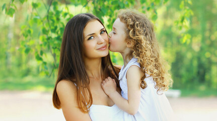 Fototapeta premium Portrait of little child daughter kissing her happy smiling mother on the grass in a summer day