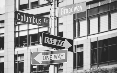 Black and white picture of a post with Columbus Circle, Broadway and one way street signs, selective focus, New York City, USA.