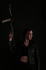 Young brunette woman in leather jacket with vintage submachine gun in darkness