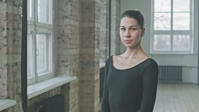 Tilt up medium portrait of young professional ballerina in black bodysuit standing alone in spacious ballet class posing to camera