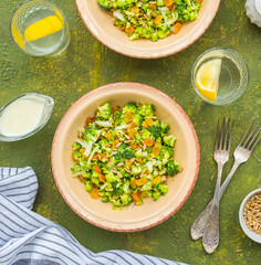 A healthy salad of raw broccoli, onions, raisins and fried sunflower seeds with yoghurt dressing in a clay plate on a green concrete background. Salad recipes, vegan food.
