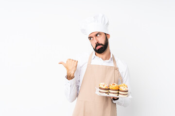 Young man holding muffin cake over isolated white background unhappy and pointing to the side