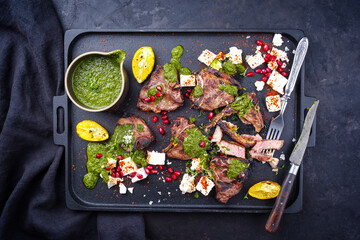 Traditional barbecue T-Bone lamb steaks with Greek feta cheese, chimichurri sauce and pomegranate fruit served as top view on a design black tray