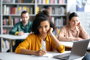 Students at lecture. Concentrated serious african american female student sitting at table in university library, preparing for lessons or exam, looks at camera, defocused students on background