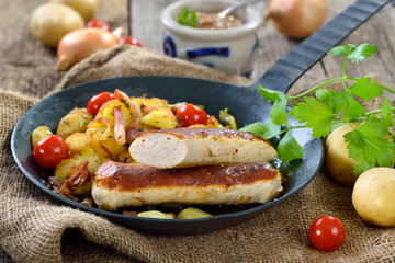 Gebratene bayerische Wollwürste mit Bratkartoffeln in der Eisenpfanne serviert – Roasted Bavarian sausages on fried potatoes with bacon, served in an iron frying pan 