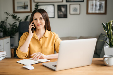 Smiling business woman talking on a mobile phone and using a laptop while working at home, looking at the screen, reading a message, a young female employee advises a client, telephone conversations