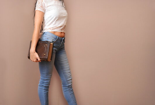 Woman in blue jeans and short white shirt stay next to the wall with large leather book in hand 