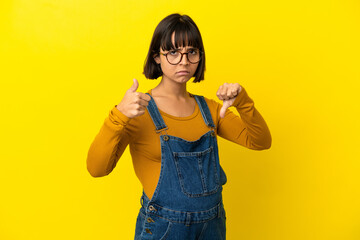 Young pregnant woman over isolated yellow background making good-bad sign. Undecided between yes or not