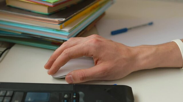 Close-up of a male hand using a white wireless computer mouse. Laptop keyboard and books on the table. A hand-held pointing device that detects two-dimensional motion relative to a surface.