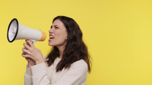 Irritated Young woman screaming in megaphone wearing basic light sweat shirt posing isolated on yellow background studio. Brunette hair Caucasian girl screaming in megaphone. slow motion. Zoom out.