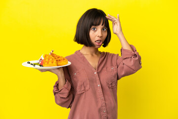 Pregnant woman holding waffles isolated on yellow background doing surprise gesture while looking to the side