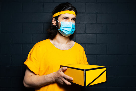 Portrait of young delivery man holding a yellow box, wearing medical mask and orange shirt on background of black brick wall.
