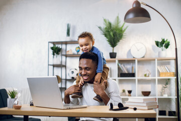Happy african father holding his cute son on neck while working on wireless laptop at home. Businessman solving working issues online using modern technology.