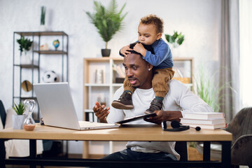 Busy afro american man working on laptop while sitting at table with son on neck. Father freelancer using portable computer for distance work.