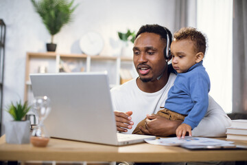 Afro american father in headset using modern laptop for video conference while holding baby boy on...