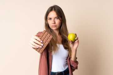 Young Lithuanian woman isolated on beige background taking a chocolate tablet in one hand and an...