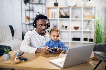Afro american father and cute son using modern laptop and wireless joysticks for playing online games. Concept of entertainment, family and leisure time.