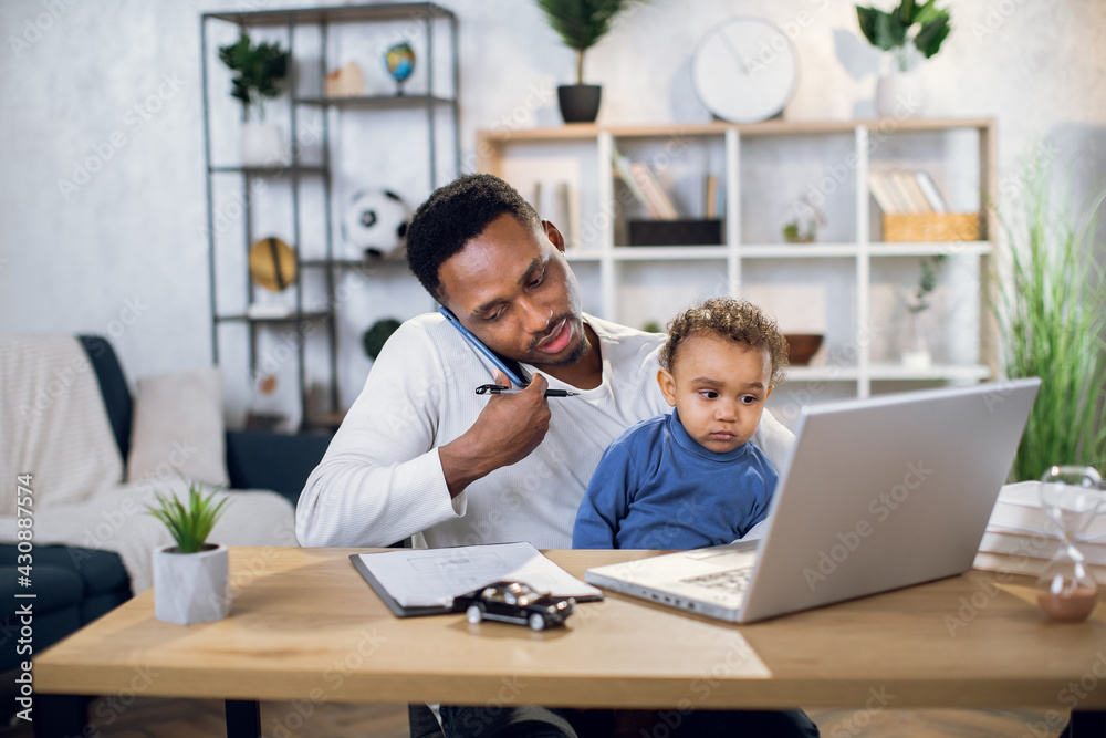 Wall mural african businessman holding his cute son on knees while sitting at table with modern laptop and talk