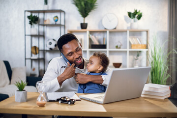 Afro american man talking on mobile, working on laptop and carrying baby son. Male freelancer...