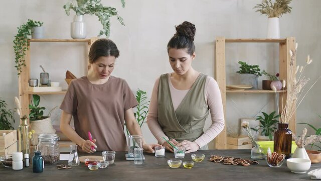 Medium shot of two attractive women practicing making homemade cosmetics. One woman telling the other ratios and proportions looking at recipe, adding bulk ingredients in measuring cup