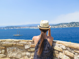 Back view of a young woman posing with a panama hat and admiring the Mediterranean sea from the...