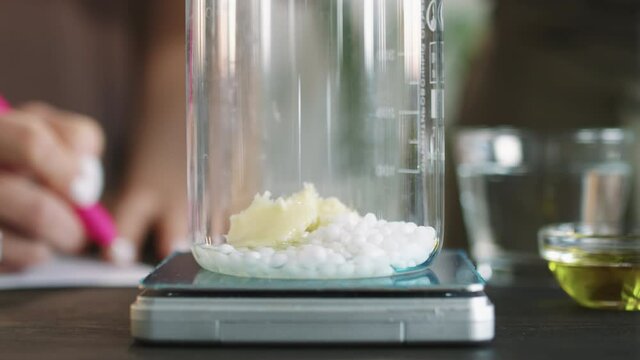 Close up of adding shea butter and small amount of oils to measuring cup with bee wax beads for making handmade face or hand cream at home