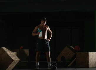 Fototapeta na wymiar Dark contrast picture of handsome young bodybuilder drinking water from a bottle and taking a break from exercising