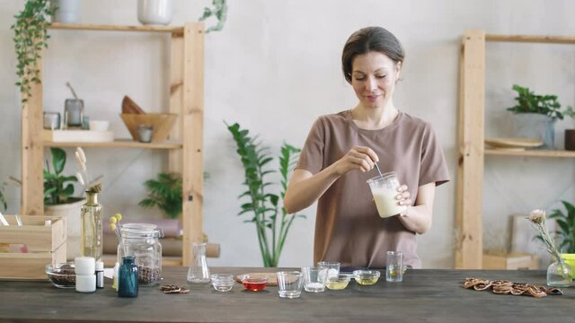 Medium PAN shot of smiling caucasian woman standing at wooden table with cup of moisturizing handmade body lotion made of shea butter, bee wax and essentials oils