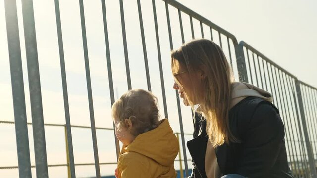 mother in black leather jacket and daughter in yellow bright raincoat look out over sea through grate or railing. Mom points her finger at something to her little girl ocean.