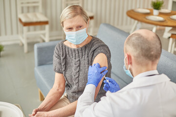 Fototapeta premium Man doctor vaccinates an elderly woman at her home wearing a protective mask. Vaccination concept
