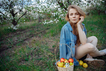 The blonde sits on a green wild lawn. There is a basket of apples next to it. The woman looks into the frame. Blooming gardens in the background. High quality photo