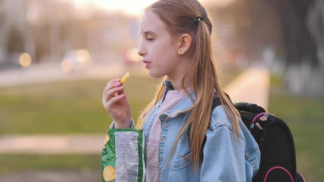 Young school girl eating chips from packet on street. Child eating chips close-up face. Little girl resting on a picnic in the park eating potato chips. Lifestyle concept.