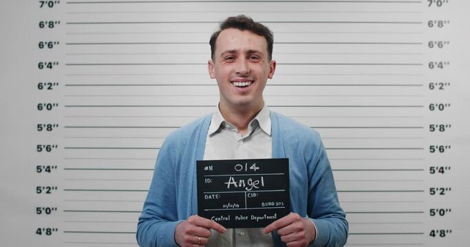 Portrait of positive man in shirt and cardigan holding sign for photo in police department. Male person in 30s posing, laughing and looking to camera. Concept of mugshot and crime.
