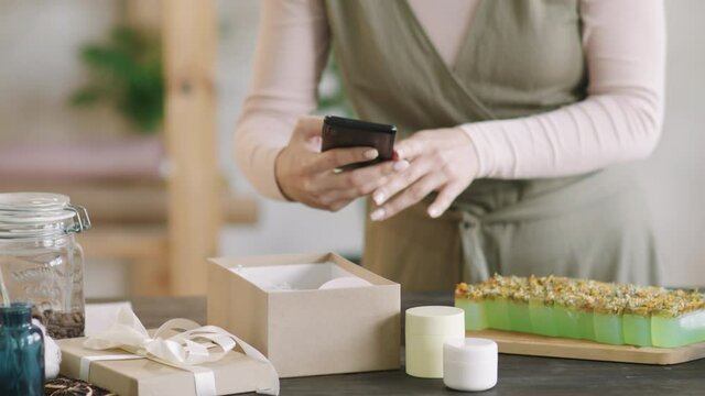 Midsection PAN shot of unrecognizable woman taking photos of handmade natural cosmetics – creams and soap – on her smartphone before posting