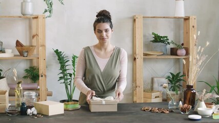 Medium PAN shot of young attractive woman packing handmade cosmetic products – face cream and soap – into beautiful present box then looking at camera smiling