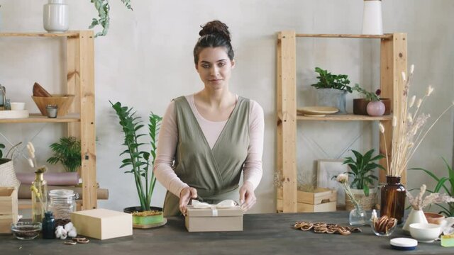 Medium PAN shot of young attractive woman packing handmade cosmetic products &ndash; face cream and soap &ndash; into beautiful present box then looking at camera smiling
