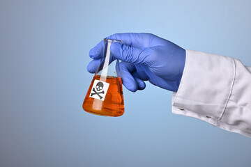 Naklejka premium Doctor hand shows toxic orange fluid in a chemical flask. Medic hand wearing a blue nitrile glove holds glass retort with orange liquid poison. Scientific research to distinct death waste or remedy.