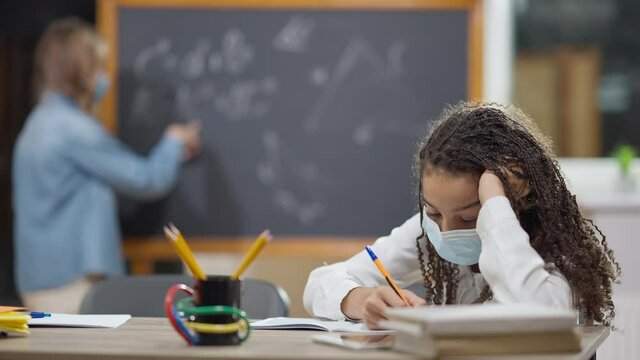 Portrait of bored African American schoolgirl in Covid face mask sitting at desk in classroom with enthusiastic Caucasian classmate writing on chalkboard at background. Sad schoolgirl studying indoors